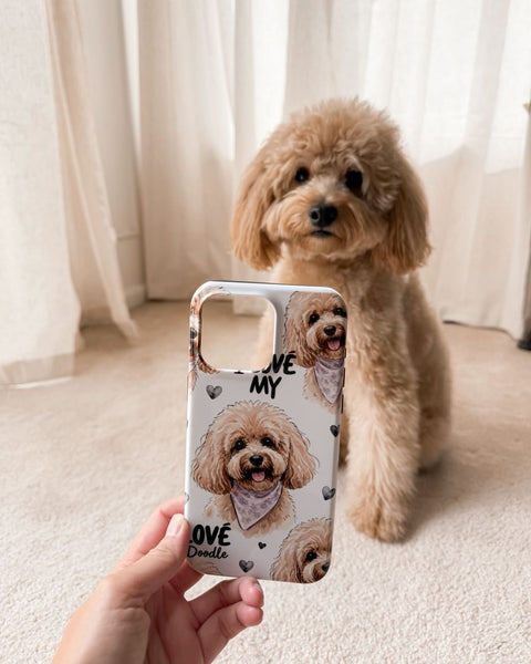 A hand holds a phone case featuring a cute cartoon doodle dog design in front of a real environment with a fluffy tan poodle and cream curtains in the background.