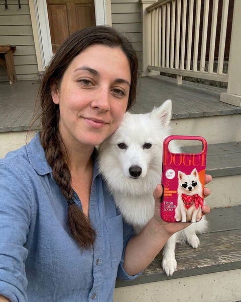 A woman on a porch holds up a pink phone case featuring a white dog illustration and the word 'DOG' in large letters, with a white dog sitting beside her.